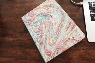 a rainbow paint pour soft cover notebook diary next to a macbook keyboard plugged into the charger