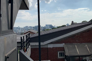 In the foreground is a residential brick house. To the left are concrete stairs. High-rise buildings stand in the distance against the backdrop of a blue sky, with some clouds within it.