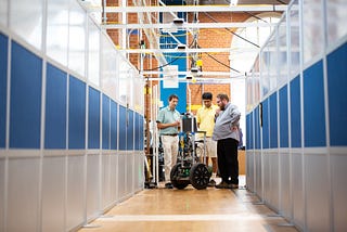 Three men stand around a robot on wheels in a robotic labs on the University of Texas at Austin campus.