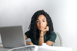 An African-American woman sitting in front of a laptop but staring at the camera with a serious expression on her face