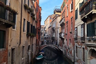 Photo of a narrow Venice canal and bridge with red, white and yellow houses on either side and a boat moored in the foreground.