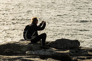 Photograph of a man with a backpack, mostly silhouetted, sitting on a rock with the sea in the background, taking a photograph on his phone.