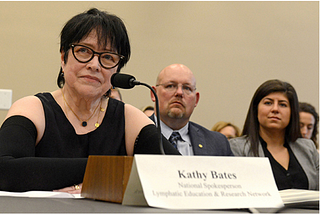 Kathy Bates with short dark hair, wearing glasses, a black top and black compression sleeves, sitting at a table with a microphone testifying before Congress