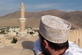 In the foreground, a man is looking at a minaret of a town surrounded by mountains in the background.