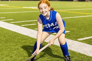 A little red-headed girl, age 11, in a blue field hockey uniform, posing for a picture on the field.