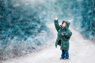 A child standing on a dirt road catches a colorful leaf falling with the snow.