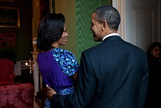Barack and Michelle Obama are standing close together and talking in the White House Green Room. Michelle wears a patterned skirt and a dark top.  Barack wears a dark suit.