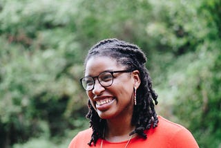 Mulher negra sorrindo, de tranças, com um óculos de grau preto.