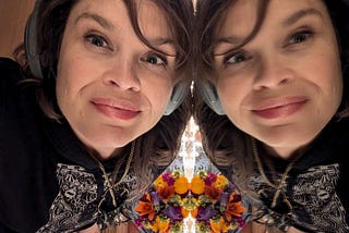 Ariel, a white woman in her late ’40s, leans her face against a mirror. She and her reflection are both looking into the lens of the camera and smiling. There are flowers in the background.