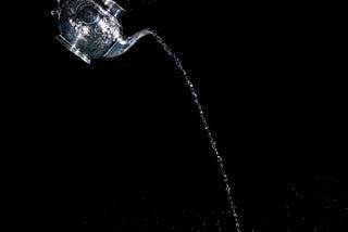 Water pouring into a silver teapot, which is in turn pouring water out of its spout. The teapot is tilted downward in mid-air on a black background.