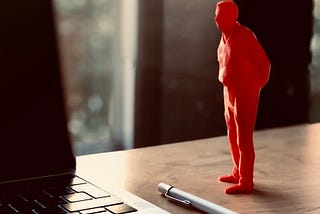 A 15 centimeter tall red plastic figurine of man with hands clasped on back “watching” your work without helping, placed on work desk by laptop.