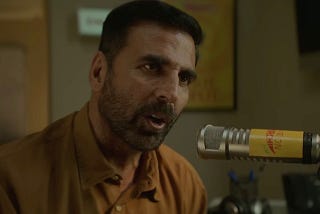 A still from Hindi film, ‘Sarfira (2024)’. A mid-length shot of Vir Mhatre (Akshay Kumar) wearing a stubble and a brown collared shirt talking into a radio mic.