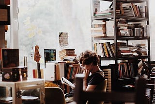 Photo of woman sitting and reading, in a room full of books (Photo by Hatice Hüma Yardım on Unsplash)
