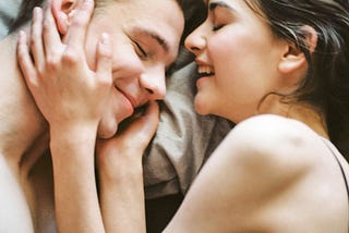 A man and female happily laying side by side in bed bolstered by their love.