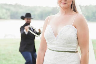A bride is at the forefront of the photo, her face showing bewilderment. Her cowboy groom is blurred in the background. He has a lasso around her waist and is about to reel her in.
