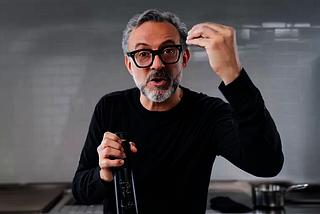 A person with short salt-and-pepper gray hair + matching facial hair and glasses talking, holding a glass bottle.