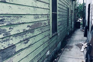 Photo of a concrete path between two wooden buildings. The side view shows several windows and paint chipping off the building on the left.