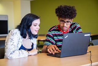 Two young people sitting at a table look at a laptop together.
