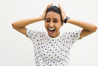 A feminine-presenting, brown-skinned person in a patterned white shirt with blue flowers and dots, has both hands on either side of their head. Their mouth is open, showing their teeth, and one eye is closed in an expression of distress. They have a nose ring on their left nostril, a silver bracelet on their right wrist, and a black watch on their left wrist. Their black hair is slicked back towards what might be a ponytail.