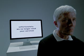A picture of a man with white hair walking away from his computer. On his screen we read the words “Unfortunately, we no longer need your services”