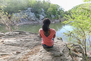 De dia, uma mulher negra está sentada sobre uma rocha, de costas para quem a observa. A frente há um rio calmo e árvores