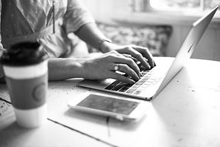 Black and white image of a women’s hands on a table typing on an Macintosh laptop. On the table there is also a coffee cup and a phone.