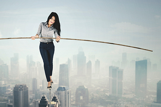 Woman with dark hair in a buttondown shirt, black slacks and high heels walking a tightrope above a smoggy city