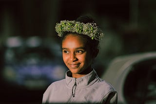 A young woman of color smiling with happy eyes, wears a crown of flowers.