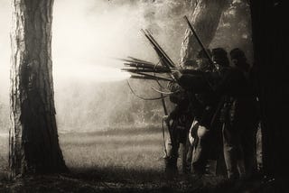 A black and white photo of Civil War soldiers in the woods, a foggy war scene playing out in a field in the background.