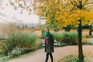 Woman in a jacket and turquoise scarf walking on a neighborhood path next to a yellow-leafed tree.