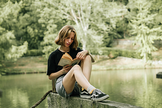 A blonde woman in a black t-shirt and shorts reads a book outside