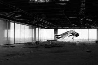 A woman floats in an abandoned, empty office space. It is a large office floor that has glass walls with a view of the city. Image is black and white.