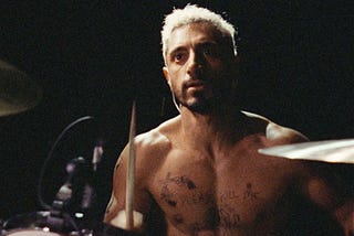 Rueben (played my Riz Ahmed) sits shirtless at his drum set in a dimly-lit room.