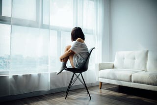 Woman in a white room sitting in a chair with feet up facing big bright window.