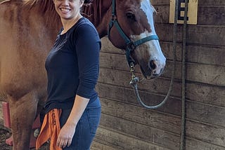 Young woman with her hair in a ponytail, wearing a blue shirt and jeans, stands smiling in front of a horse.