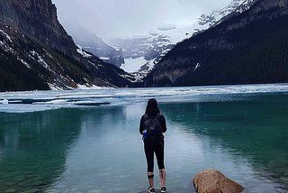 Natalie standing on a small rock facing a deep turquoise coloured partly icy Lake Louise with snowy mountains in the background.