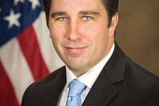 Profile for Assistant Attorney General, Stephen Boyd. He’s wearing a suit with a light blue tie; American flag in background.