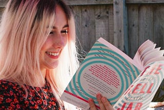 Photo of myself, sat outside in the sunshine reading a copy of The Bell Jar with a swirling aqua and pink cover.