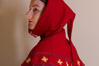 A red medieval knit hood that drapes as a collar around the shoulders with yellow four-point flowers on the border. It is being worn by a white female looking to the side against a plain white backdrop.