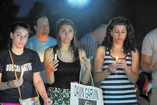 At night, three young women hold lit candles. The young woman in the middle holds a sign that says “Dawn Garvin.” Other attendees are visible behind them and to the far right.