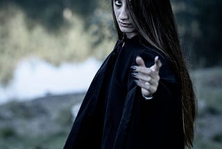 Woman with a gaunt face, long hair, black robes and long black nails reaches out one hand, in the background are trees and a lake