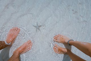 Feet of man and woman in shallow beach water with starfish between them on white sand.