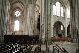 Rows of wooden pews in the foreground of an empty French gothic cathedral. Bright white light shines in through the stained glass windows. Wooden chairs in the background.