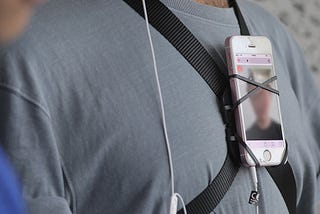 A person in a gray shirt whose phone is attached to their chest with a harness. The screen displays a blurry image of someone’s face.