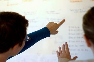 Christy pointing at a whiteboard covered in notation with a collegue