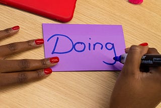 Um post-it roxo com a palavra “Doing” escrita em azul com uma caneta piloto por uma mulher negra com unhas pintadas de vermelho