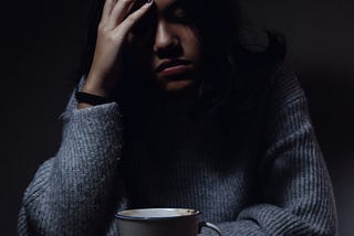 An image of a young woman in seated in a dark setting with head held against her right palm, in a depressed pose.