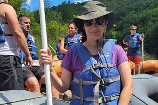 Me sitting in a inflatable raft posing with an oar before the rafting trip on a beautiful sunny day. Other people are standing around me all waiting and ready to go.