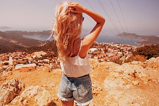 An attractive blonde woman in denim cutoff shorts, a white cotton tank top, and dark sunglasses gazes down from a sunbaked rocky hillside to a small town situated along the ocean.