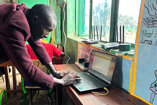 A teacher stands up using Kolibri on a laptop on the desk in front of him.